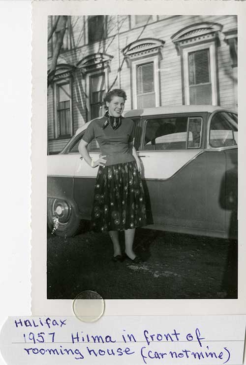Photo en noir et blanc d’une jeune femme devant une vieille voiture.