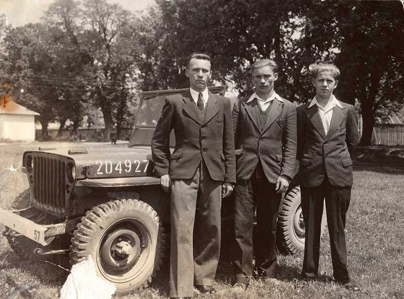 Trois jeunes hommes en costume sombre se tiennent devant une vieille jeep.