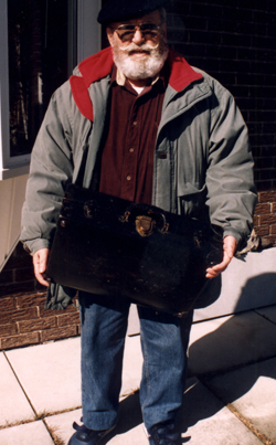 Bearded man wearing a blue beret, dragging an old suitcase.