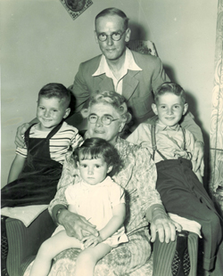 Femme plus âgée assise dans un fauteuil avec trois enfants autour d’elle et un homme debout.