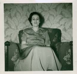 Joan en chemisier et jupe, assise sur le divan d’un salon. 