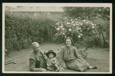 La jeune Joan portant un chapeau, assise dans l’herbe d’un jardin avec ses parents. 