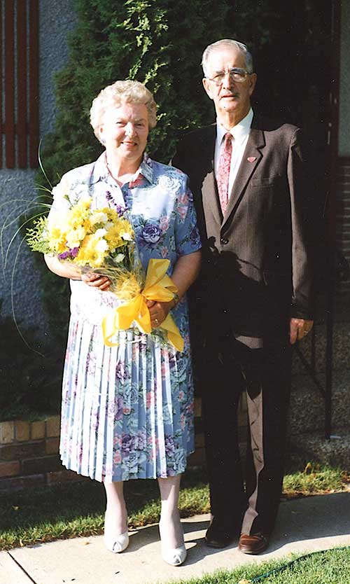 Un homme et une femme âgés se tiennent devant leur maison, la femme tient un grand bouquet jaune.