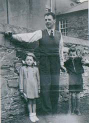 Homme adossé à un mur de pierre, un petit garçon et une fille de chaque côté.
