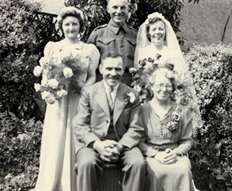 Deux jeunes belles dames portant robe de mariée tenant des fleurs debout avec bel homme et vieux couple assis en face d’eux.