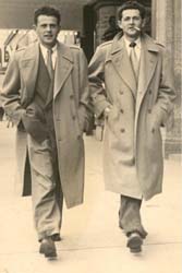 Deux jeunes hommes en costume et pardessus, marchant sur le trottoir. 