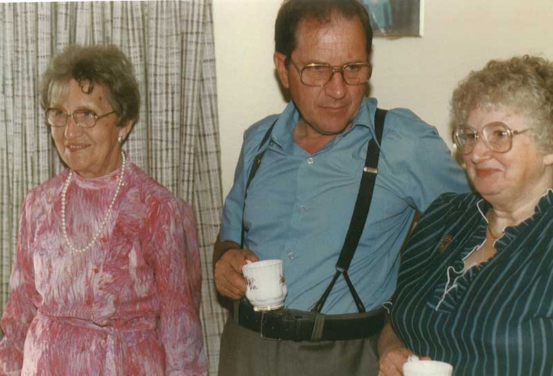 Un homme qui tient une tasse de thé et qui porte des bretelles. Il est debout entre deux femmes.