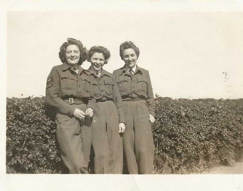 Trois femmes se tiennent devant une haie, elles portent toutes des uniformes militaires.