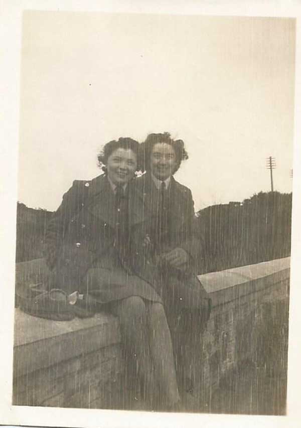 Deux femmes souriantes en tenue militaire sont assises sur un mur de briques.