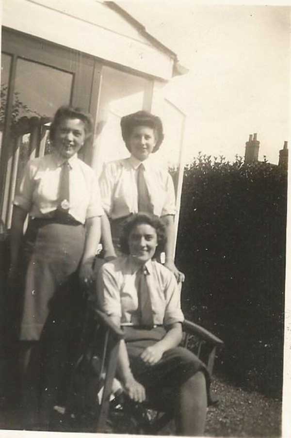 Trois femmes sont représentées, une assise et deux debout derrière elle. Elles portent toutes des chemises blanches et des cravates sombres.