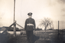 Le jeune William en uniforme, debout devant une clôture en fils barbelés.