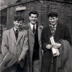 Trois jeunes hommes en costume et pardessus, debout devant un immeuble.