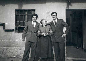 Femme plus âgée se tenant entre deux jeunes hommes, devant l’immeuble.