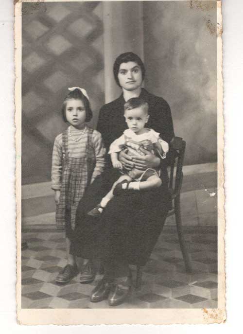 Photo archivistique d’une femme assise dans une chaise avec un tout-petit sur ses genoux et d’une jeune fille debout à côté d’elle.