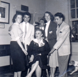 Femme en robe noire assise, avec cinq jeunes adultes autour d’elle.