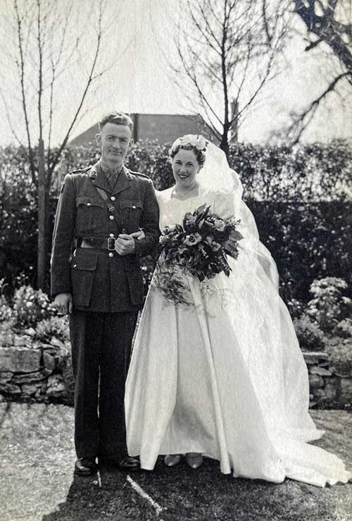 Une jeune femme porte une robe de mariée et tient des fleurs, tout en se tenant à côté d’un jeune homme en uniforme militaire.