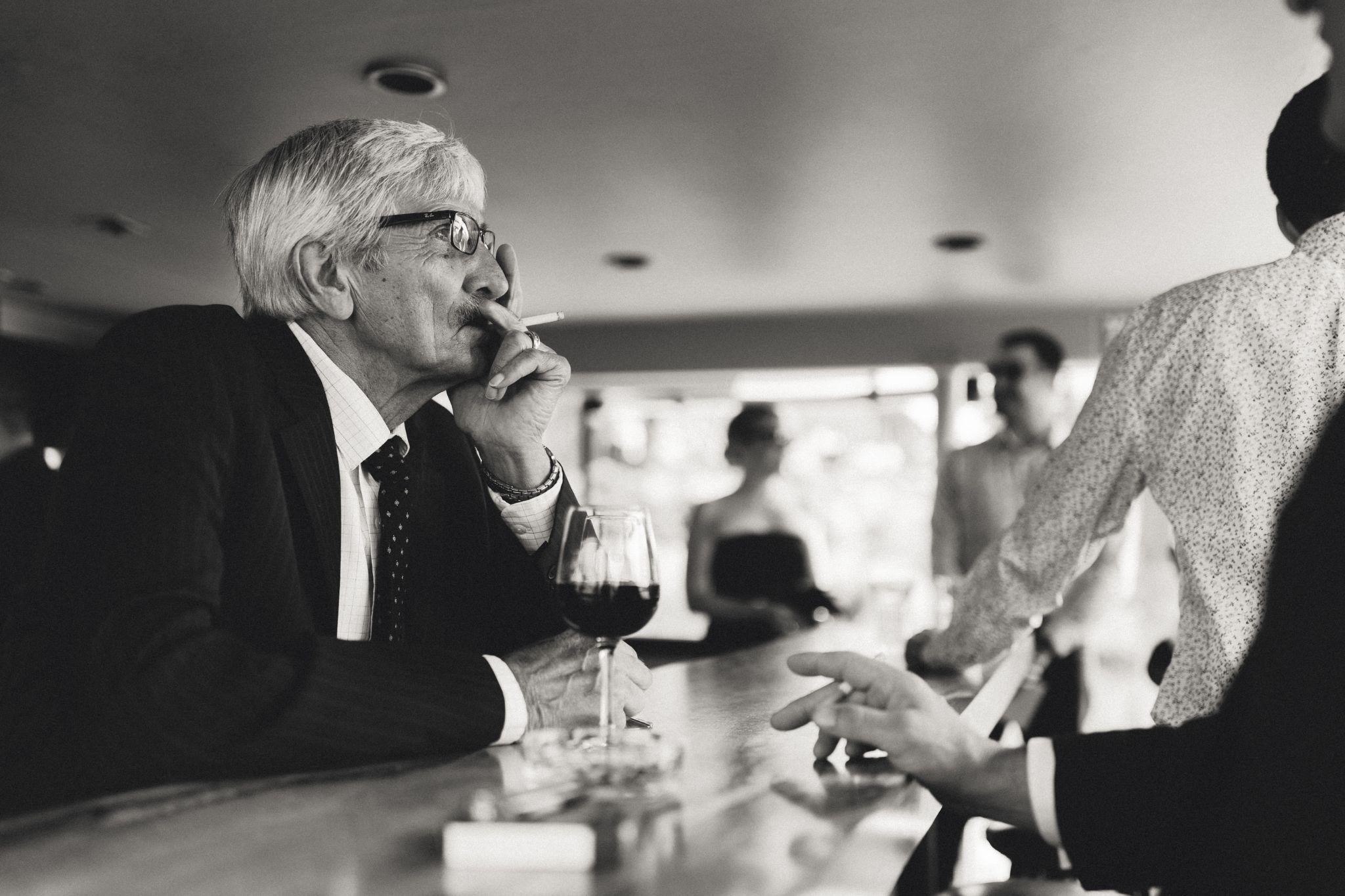 Photo en noir et blanc d’un Italien en costume fumant une cigarette dans un bar avec un verre de vin.