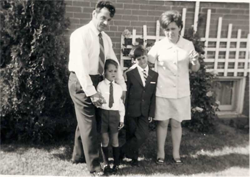 Une jeune famille pose à l’extérieur de leur maison; le père et la mère et deux jeunes garçons vêtus d’uniformes scolaires.