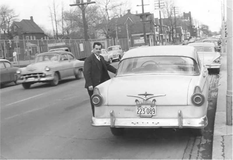 Un jeune homme se tient à côté de sa voiture en regardant la caméra.