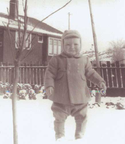 Un bébé debout sur la neige devant la maison.