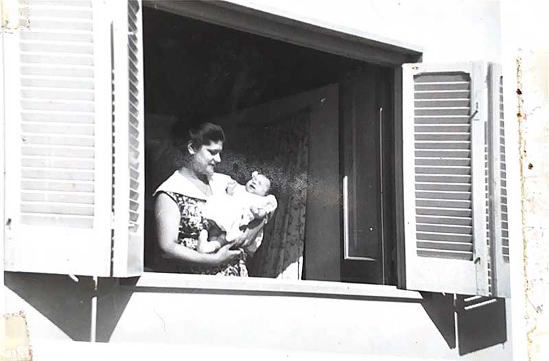 Une femme tenant un bébé se tient dans une fenêtre, les volets sont ouverts.