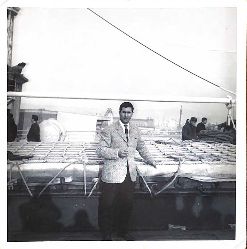 Un homme en costume est sur le pont d’un navire, il fume une cigarette.
