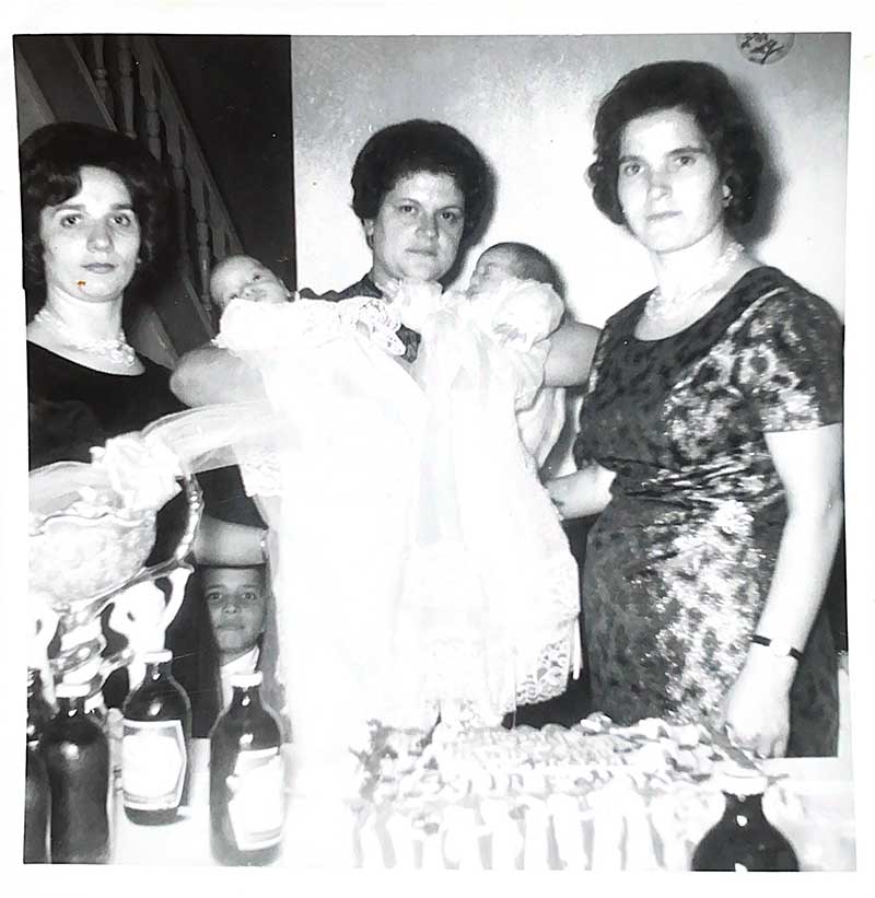 Une femme tient des bébés jumeaux dans des robes de baptême, deux femmes sont debout de chaque côté d’elle.