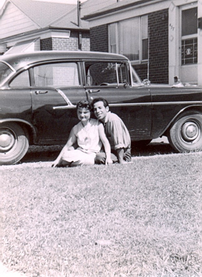 Photo en noir et blanc d’un homme et d’une femme italiens assis dans une allée avec une voiture derrière eux.