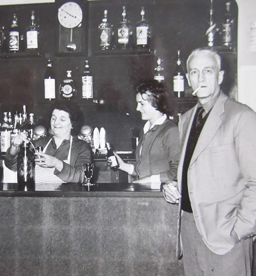 Un homme se tient devant un bar en train de fumer une cigarette, tandis que deux femmes sont derrière le bar.