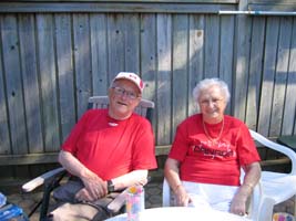 Photographie récente de Noel et de Noreen, plus âgés, portant des t-shirts rouges, assis sur le patio.
