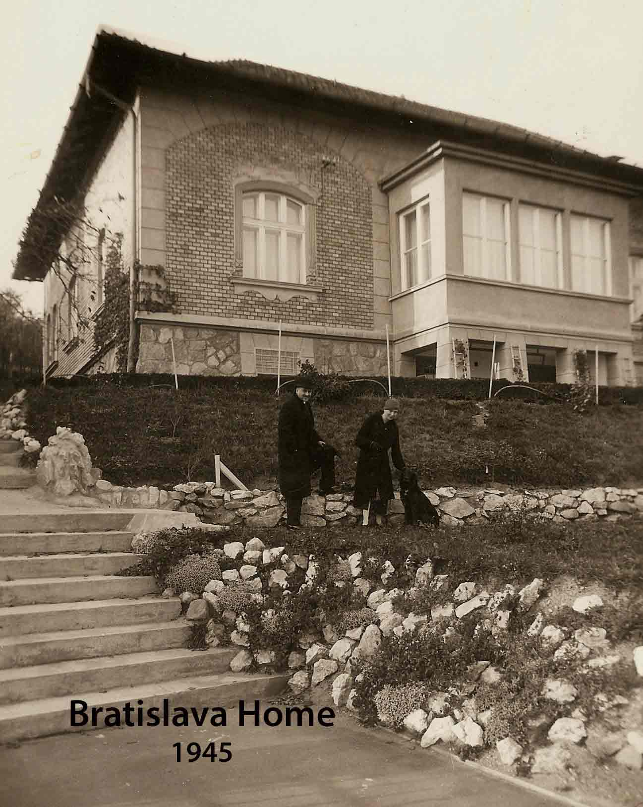 Photo en tons sépia de deux personnes et d’un chien devant une maison avec un rebord en briques.