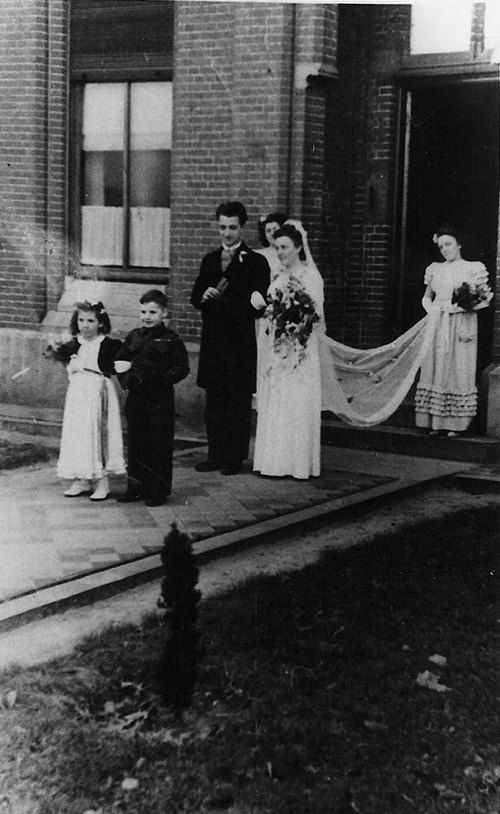 Vieille photo de couple marié sortant de l’immeuble. Deux enfants marchent devant eux et une dame tient la robe de mariée.
