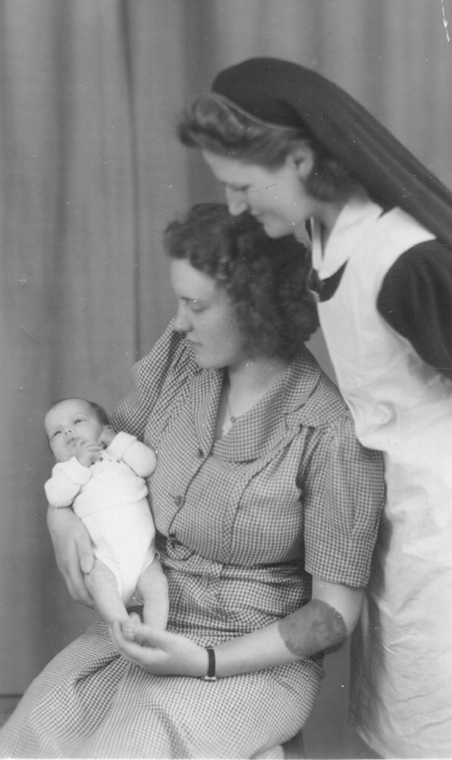 Une belle dame assise sur un tabouret tenant un nouveau-né avec une autre femme debout derrière elle.