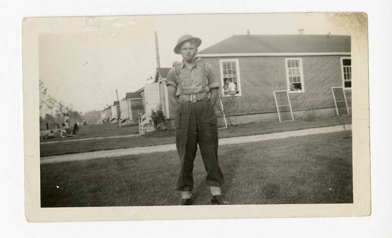 Un jeune homme en uniforme militaire se tient sur l’herbe devant une maison.