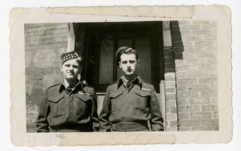 Deux jeunes hommes en uniforme militaire se tiennent devant un bâtiment en ciment.