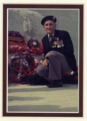 Photographie récente de Langille plus âgé, en tenue militaire, à genoux près d’une couronne de fleurs posée près d’un cénotaphe.