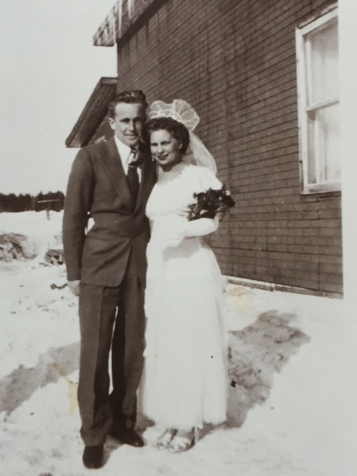 Une mariée et un marié se tiennent devant un bâtiment avec les bras l’un autour de l’autre.