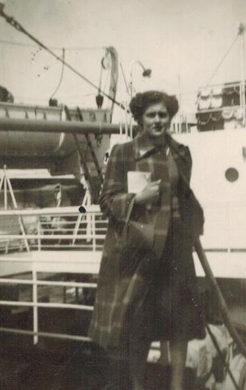 Une femme est debout sur le pont du navire portant un long manteau à damier et tenant un livre dans une main.