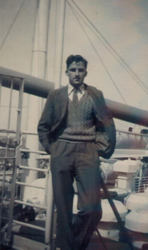 Un jeune homme portant un costume en trois pièces se penche contre la rambarde d’un navire et a les mains dans ses poches.