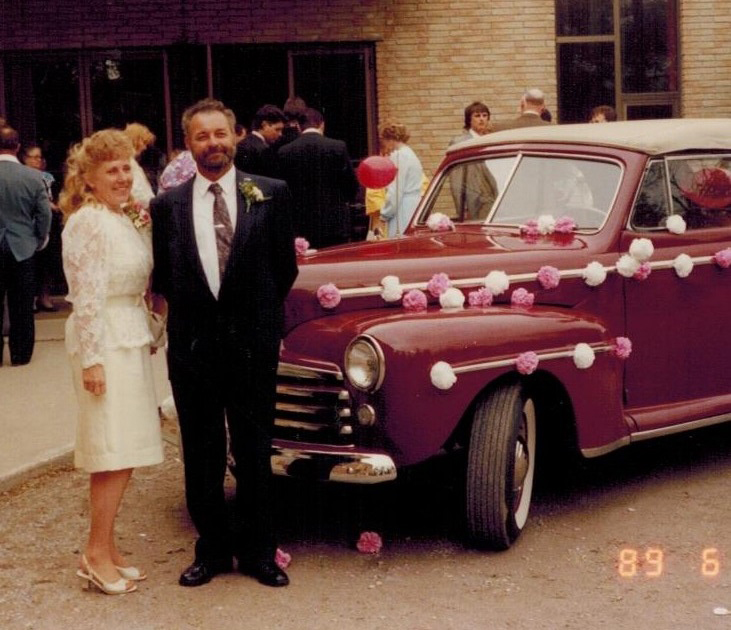 Un homme et une femme se tiennent devant une vieille voiture décorée de fleurs de papier et de banderoles.