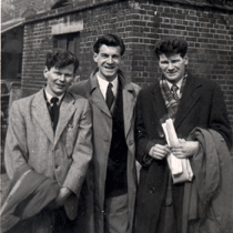 Trois jeunes hommes en pardessus devant l’immeuble.