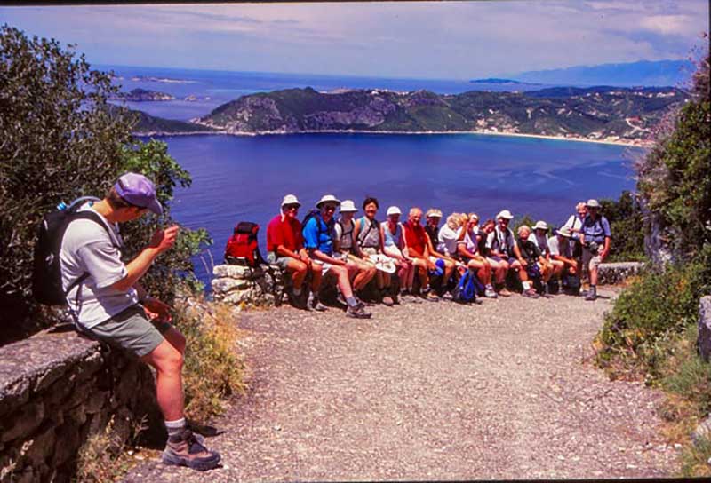 Un groupe de touristes assis dans une rangée sur un mur de roche, avec l’eau et les collines derrière eux.