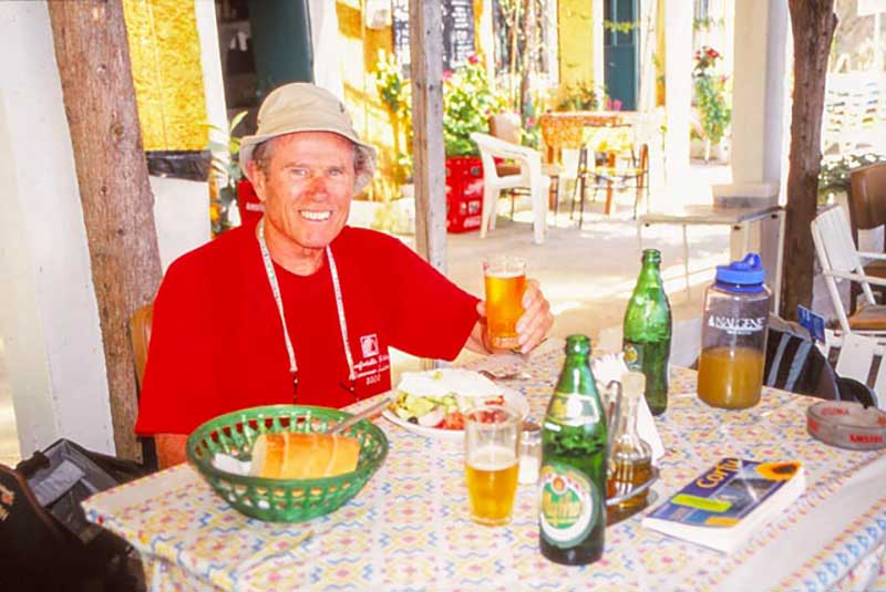 Un homme en t-shirt rouge est assis à une table remplie de nourriture et de boissons.