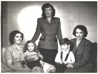 Portrait de famille avec femme debout au milieu de deux femmes assises, chacune tenant un petit enfant.