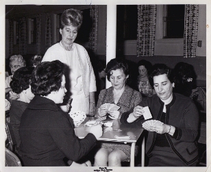 Quatre femmes assises, jouant aux cartes, une femme en blanc se tenant au-dessus. 