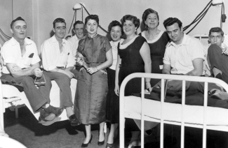 Hommes assis sur des lits d’hôpital et femmes debout à leur côté. 