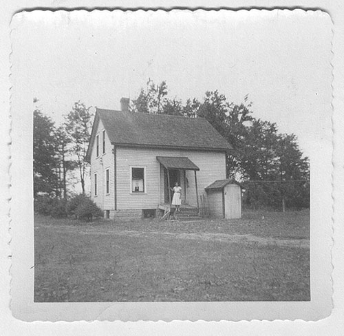 Une petite ferme au milieu d’un champ, une jeune fille en robe blanche se tient sur le porche.