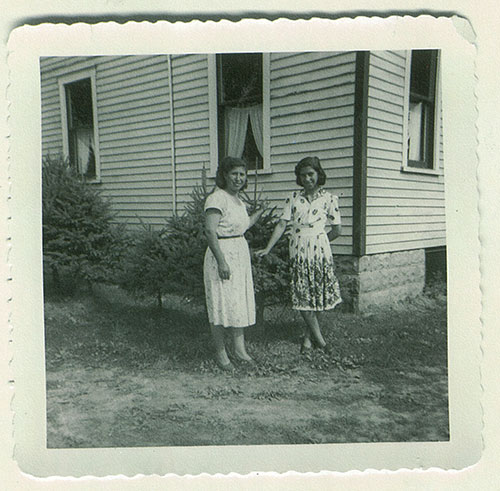 Deux femmes se tiennent devant une maison, devant un arbuste.