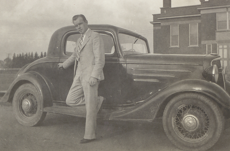 Un homme en costume et cravate se dresse contre une voiture, tenant la poignée de la porte.