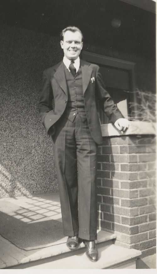 Un homme souriant dans un costume trois-pièces se tient sur les marches d’un bâtiment.